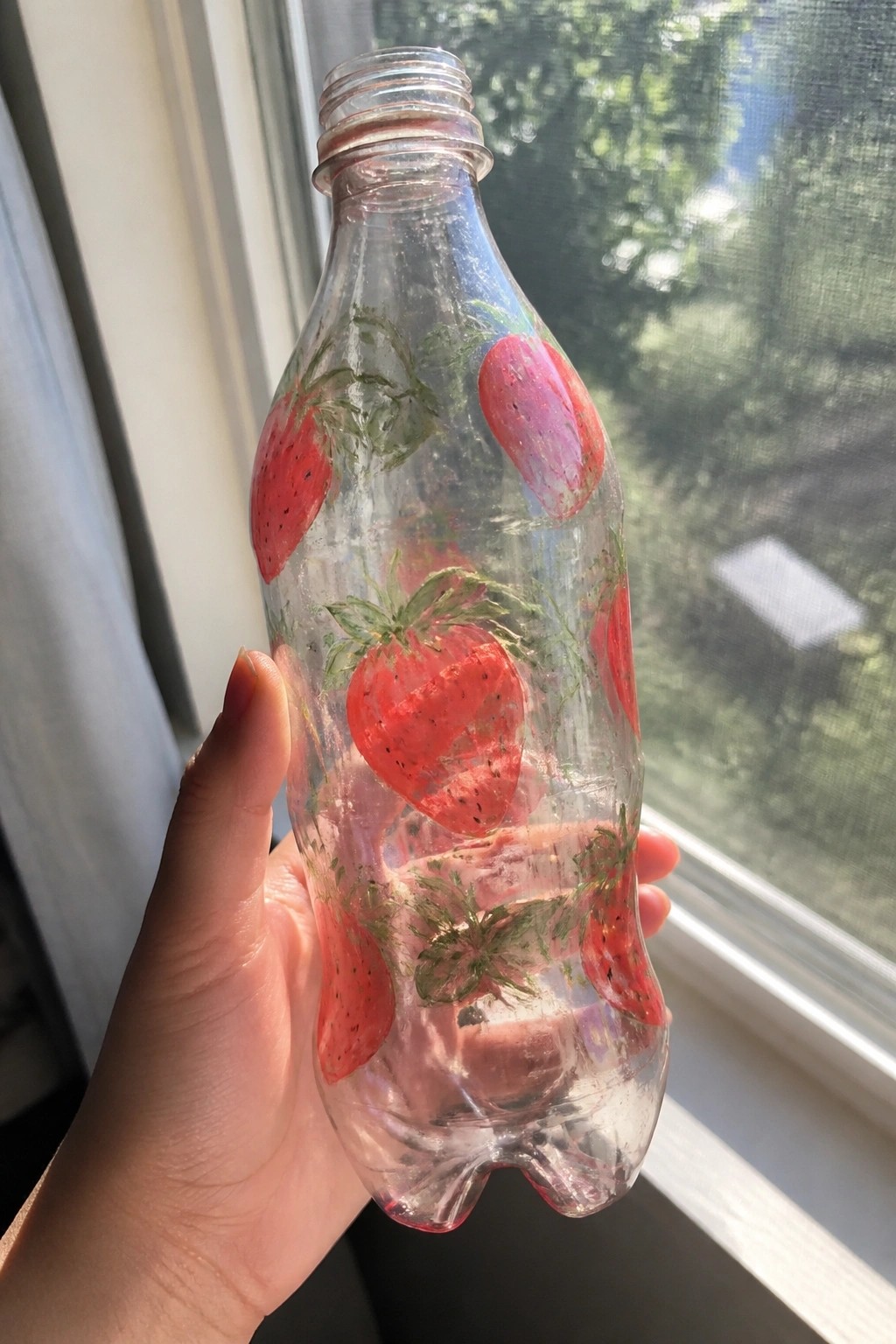 Hand holding clear plastic bottle painted with red strawberries and green leaves by window.