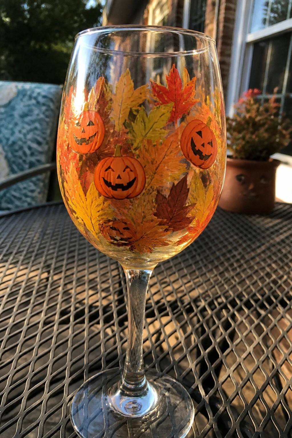 A clear wine glass with orange pumpkins and fall leaves painted on the bowl rests on an outdoor metal table.