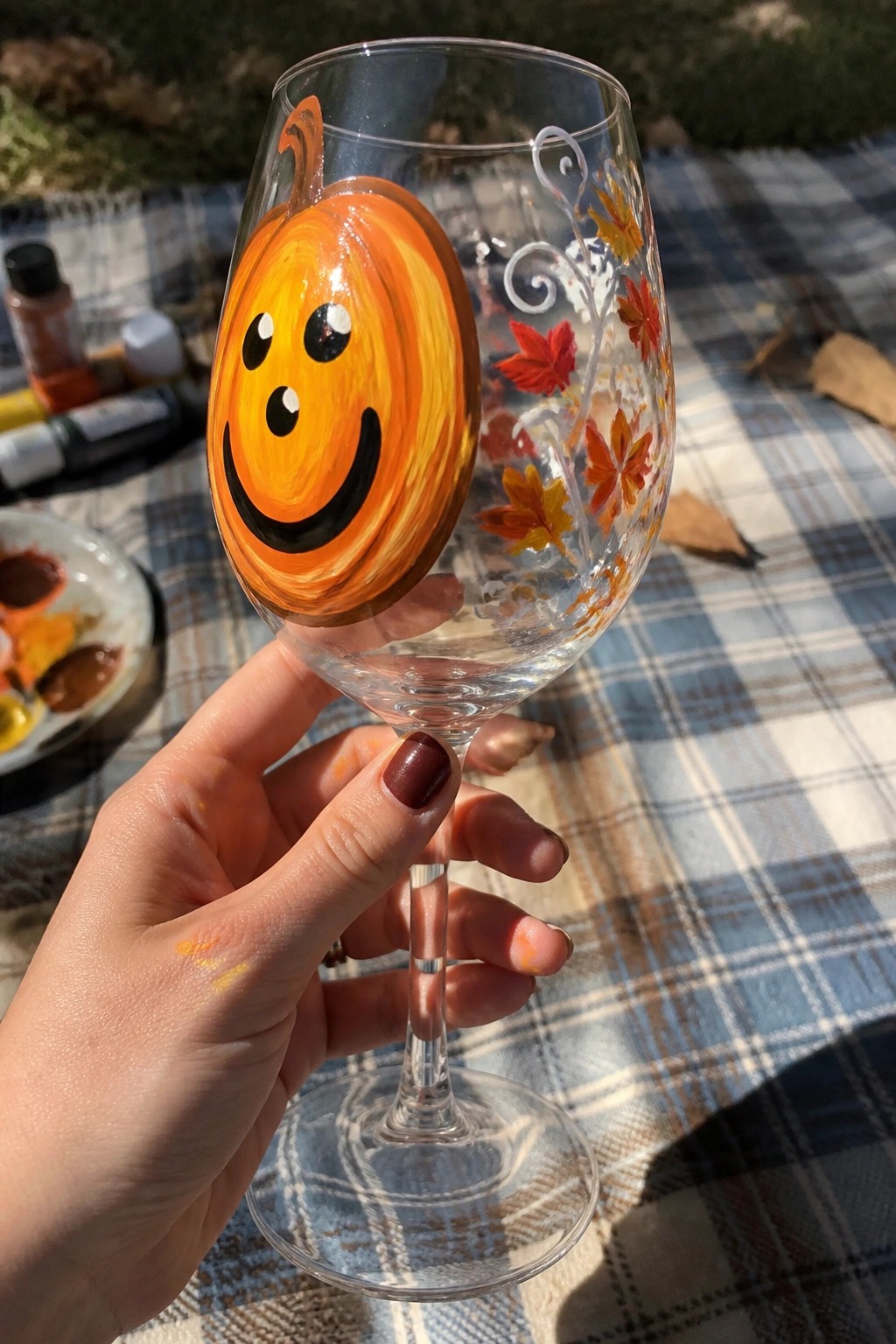 Hand holding wine glass painted with smiling pumpkin and autumn leaves on plaid blanket.
