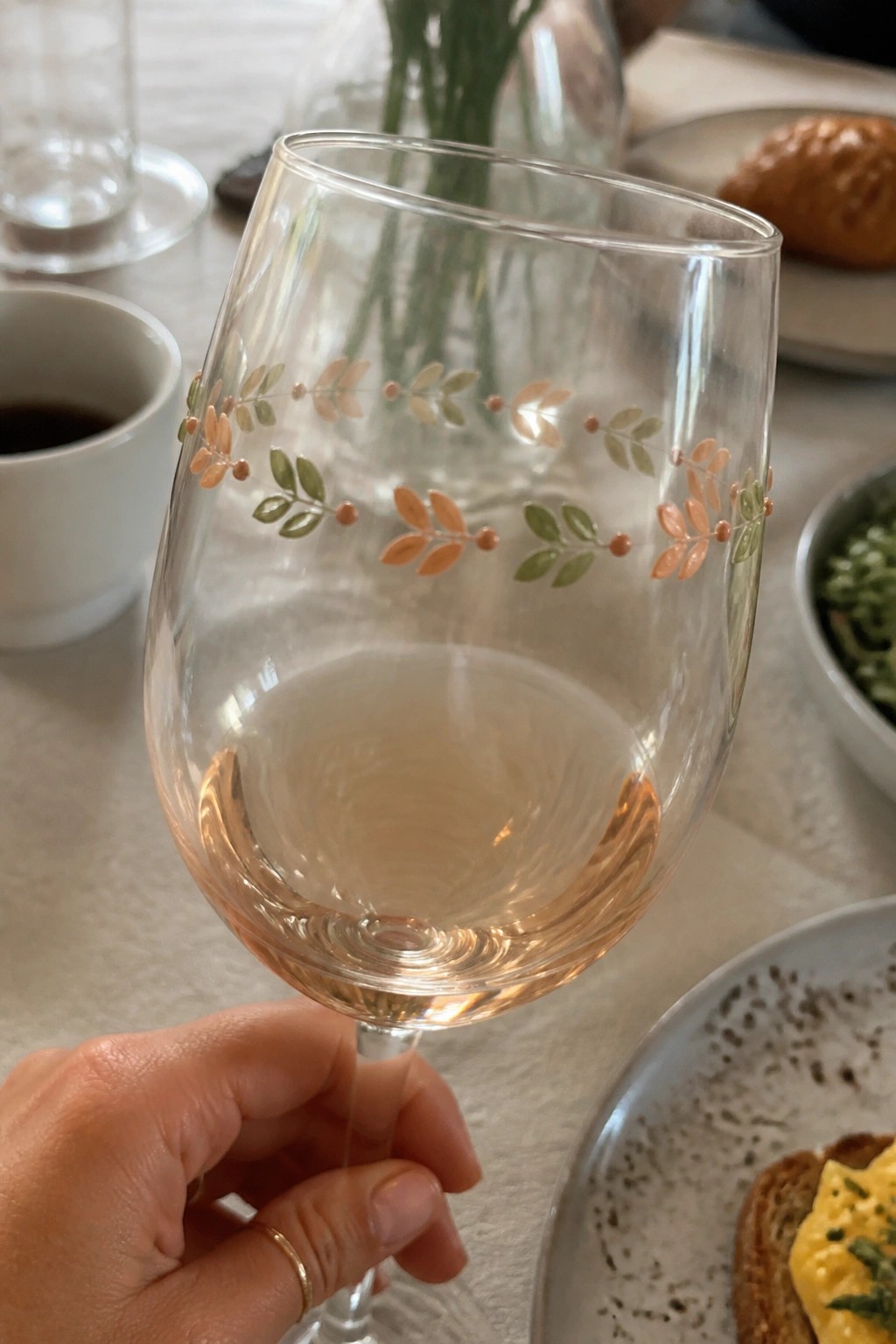 Hand with gold ring holding leaf-decorated glass of rosé at breakfast table.