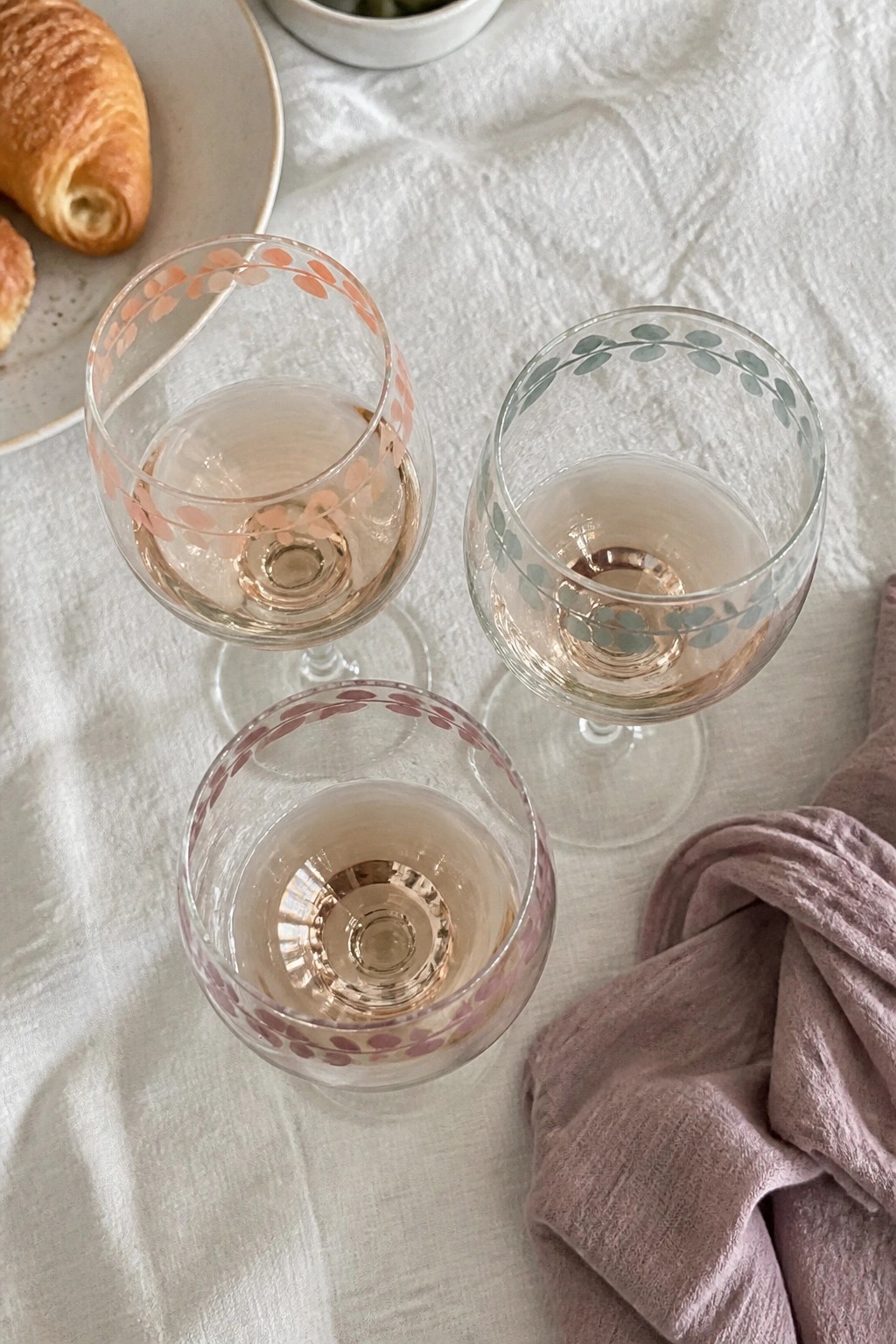 Three patterned wine glasses of rosé beside croissant and purple napkin on linen.