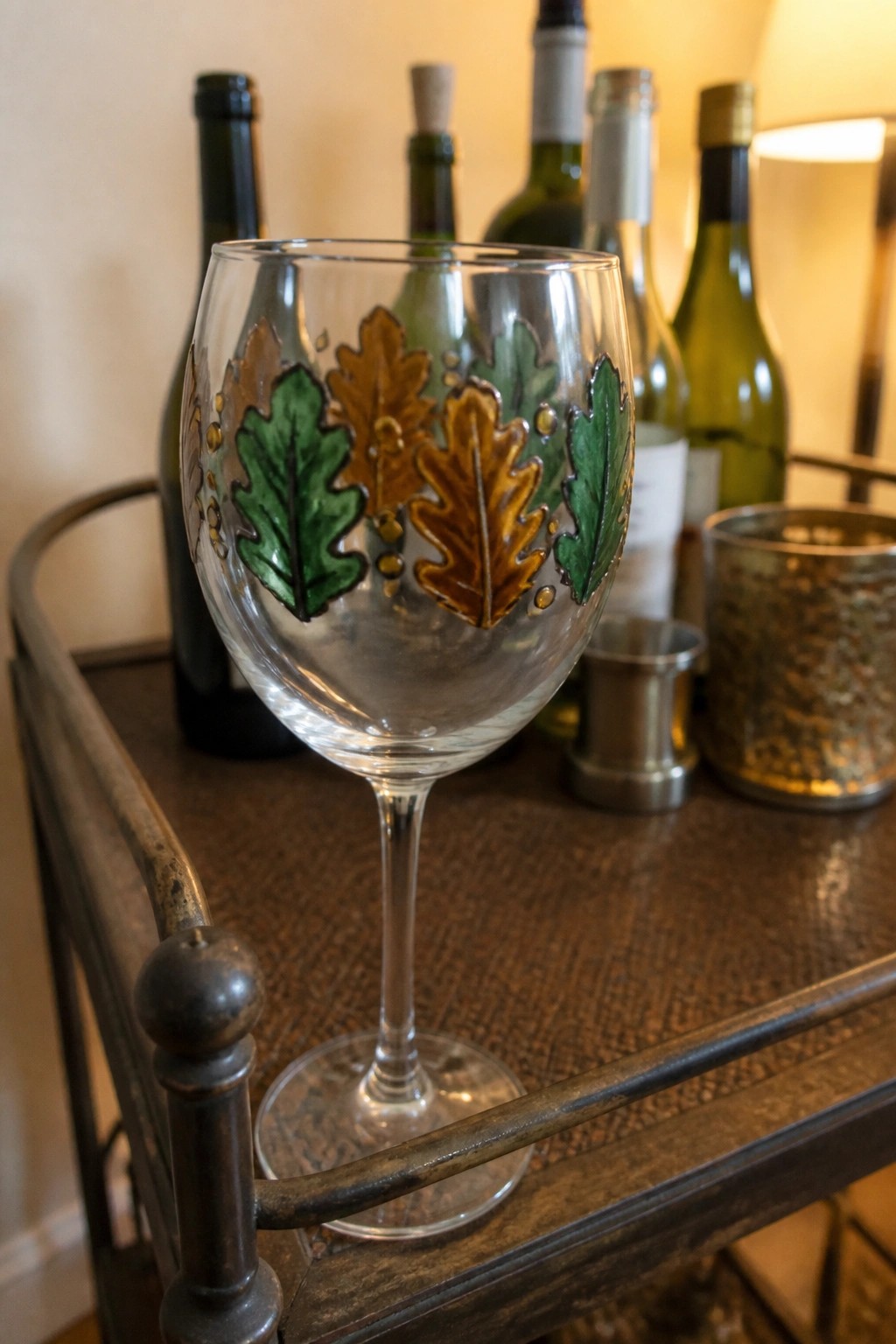 Decorated wine glass with green and brown oak leaves on wooden tray amid bottles.