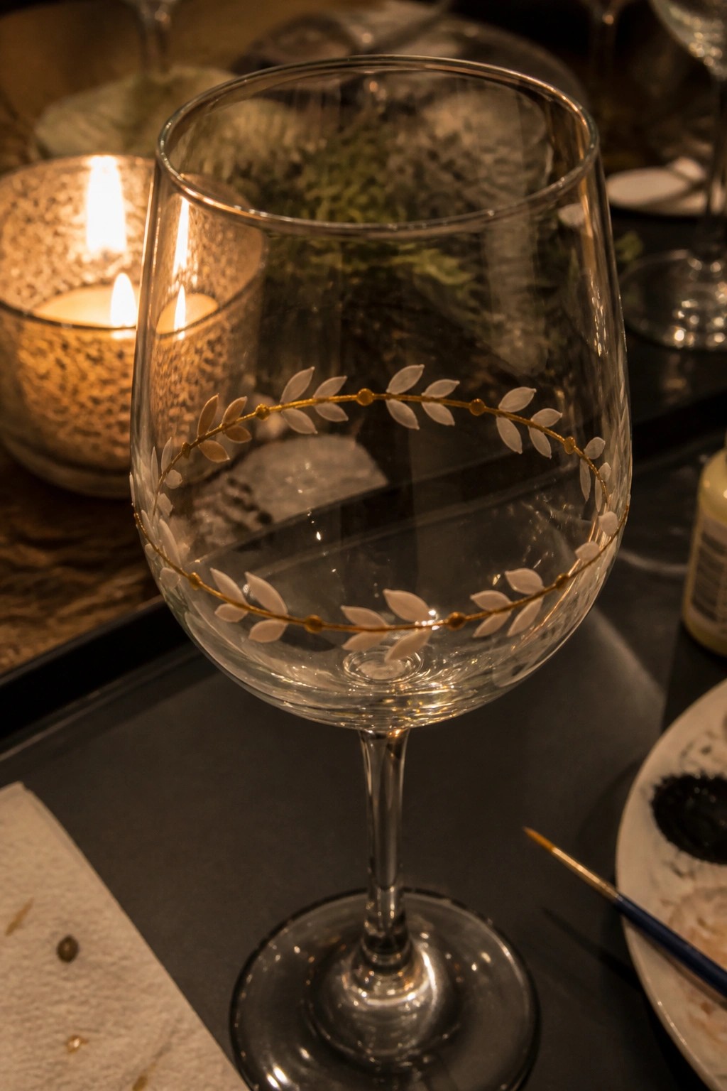 Wine glass with gold and white leaf design beside lit candle and paintbrush