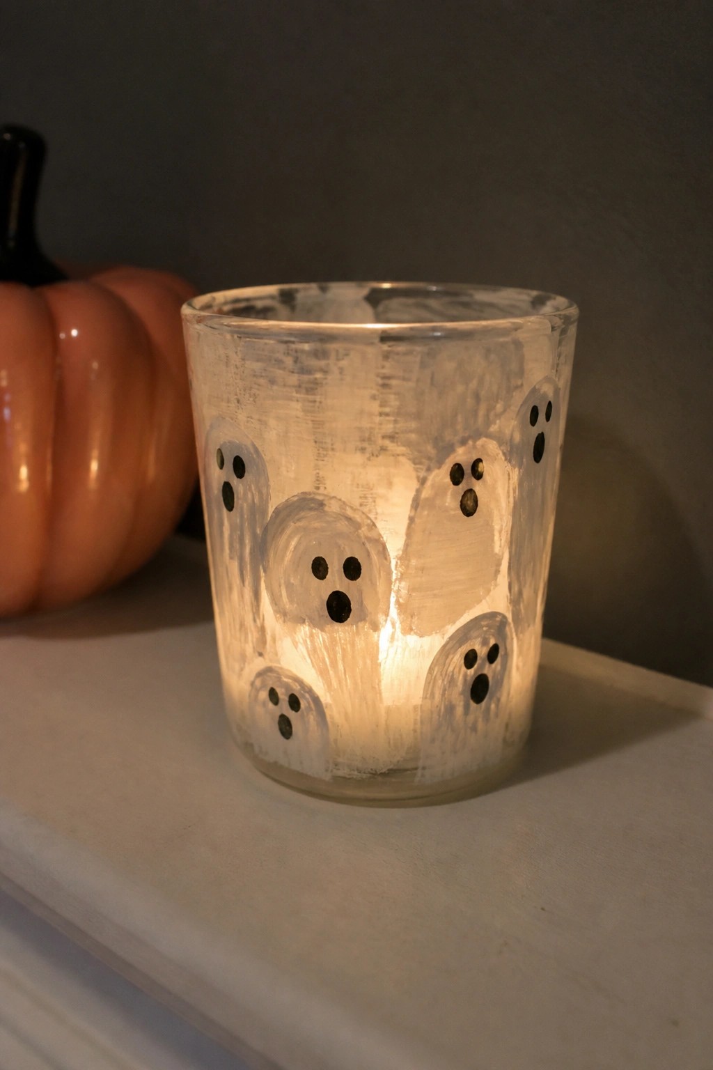 Glowing glass candle holder painted with ghost figures beside an orange pumpkin