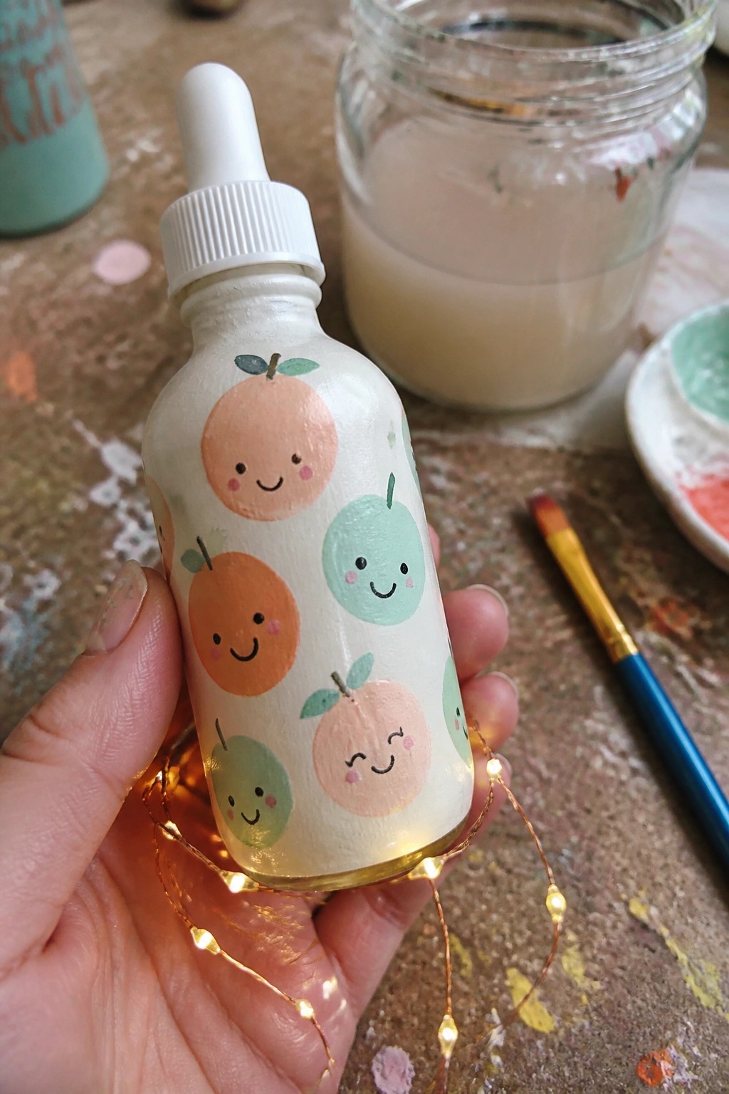 Hand holding white dropper bottle painted with cute smiling fruit faces and glowing lights