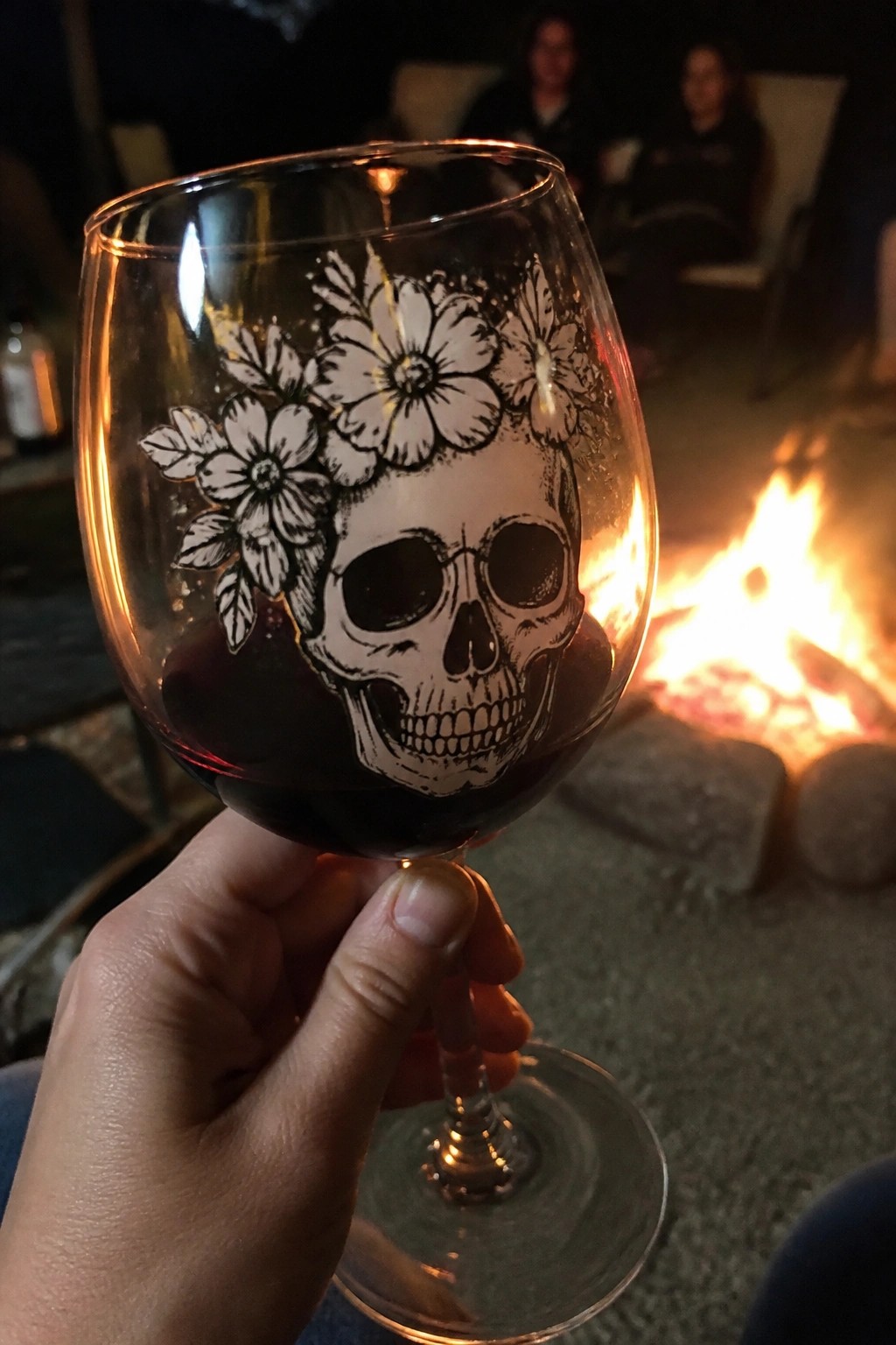 Hand holding red wine glass etched with skull and flowers by campfire at night