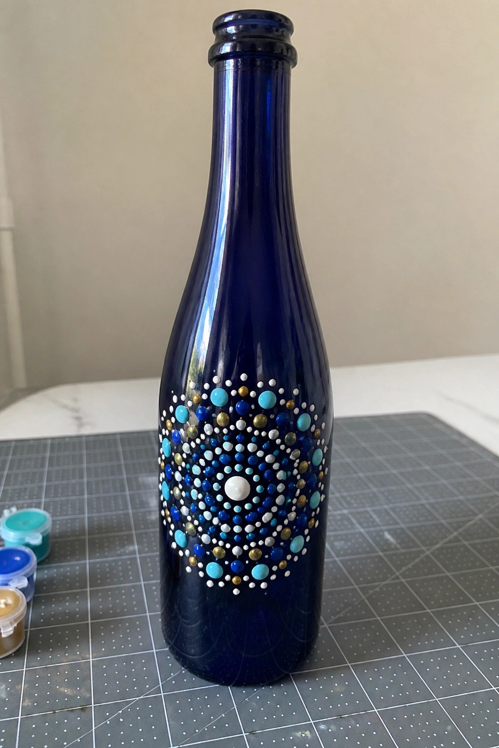 Blue glass bottle with turquoise, white, and gold dot mandala on cutting mat
