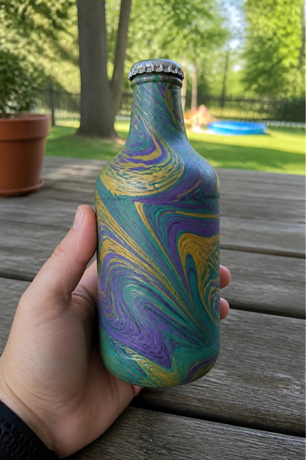 Hand holding marbled teal purple gold painted bottle on wooden deck outdoors