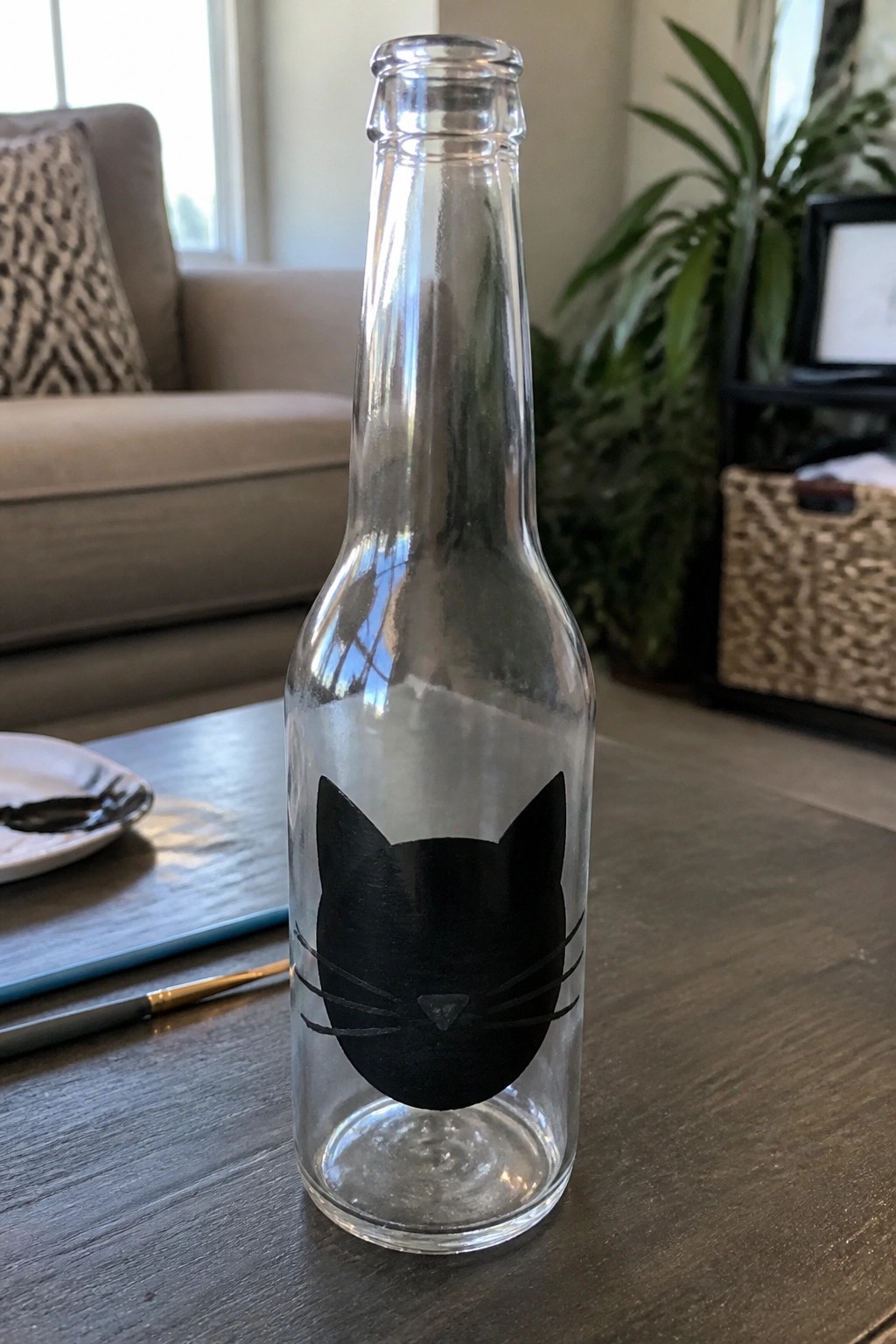 Clear glass bottle with black cat face painting on wooden table beside paintbrush