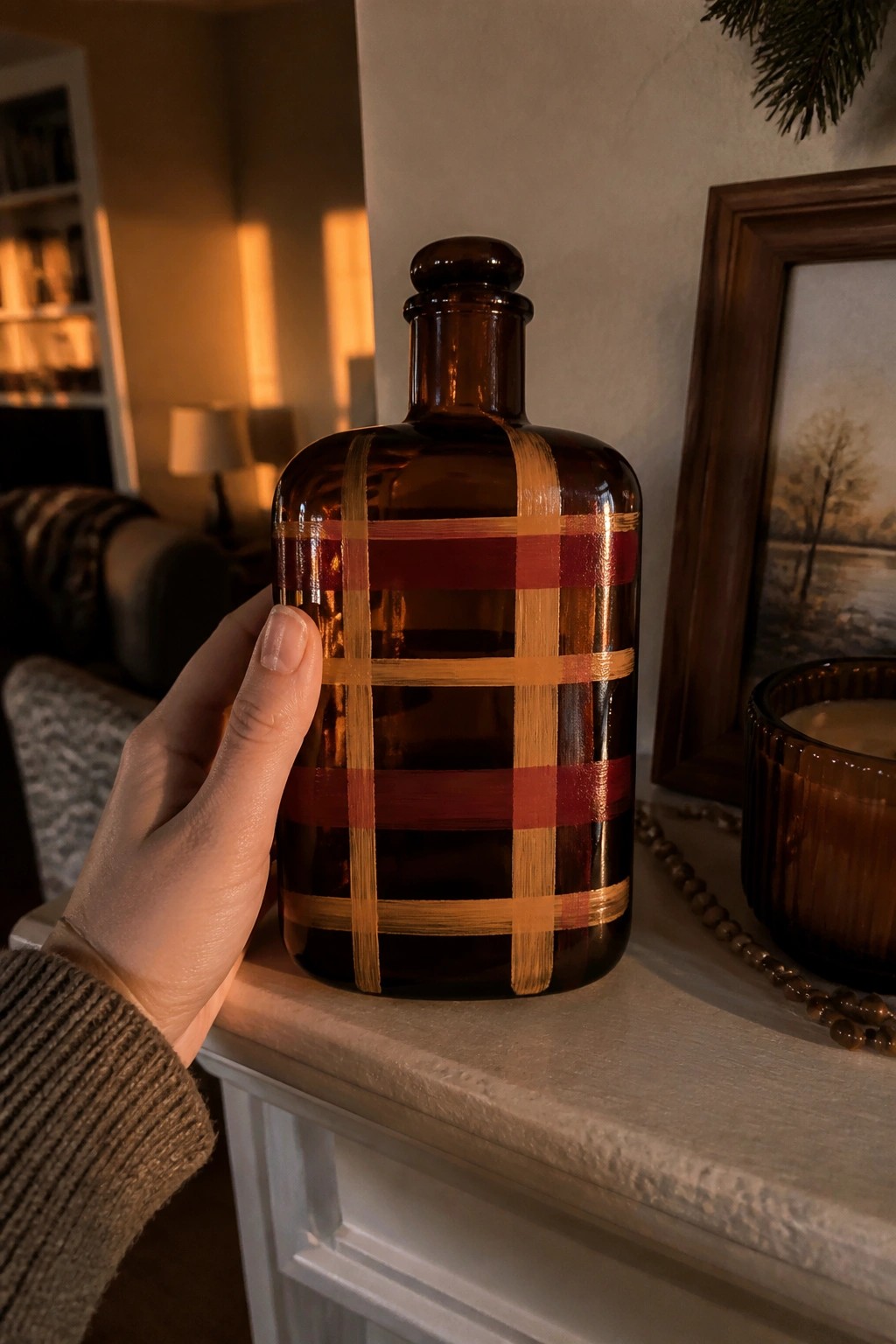 Hand in brown sweater holding plaid-painted amber glass bottle on mantel.