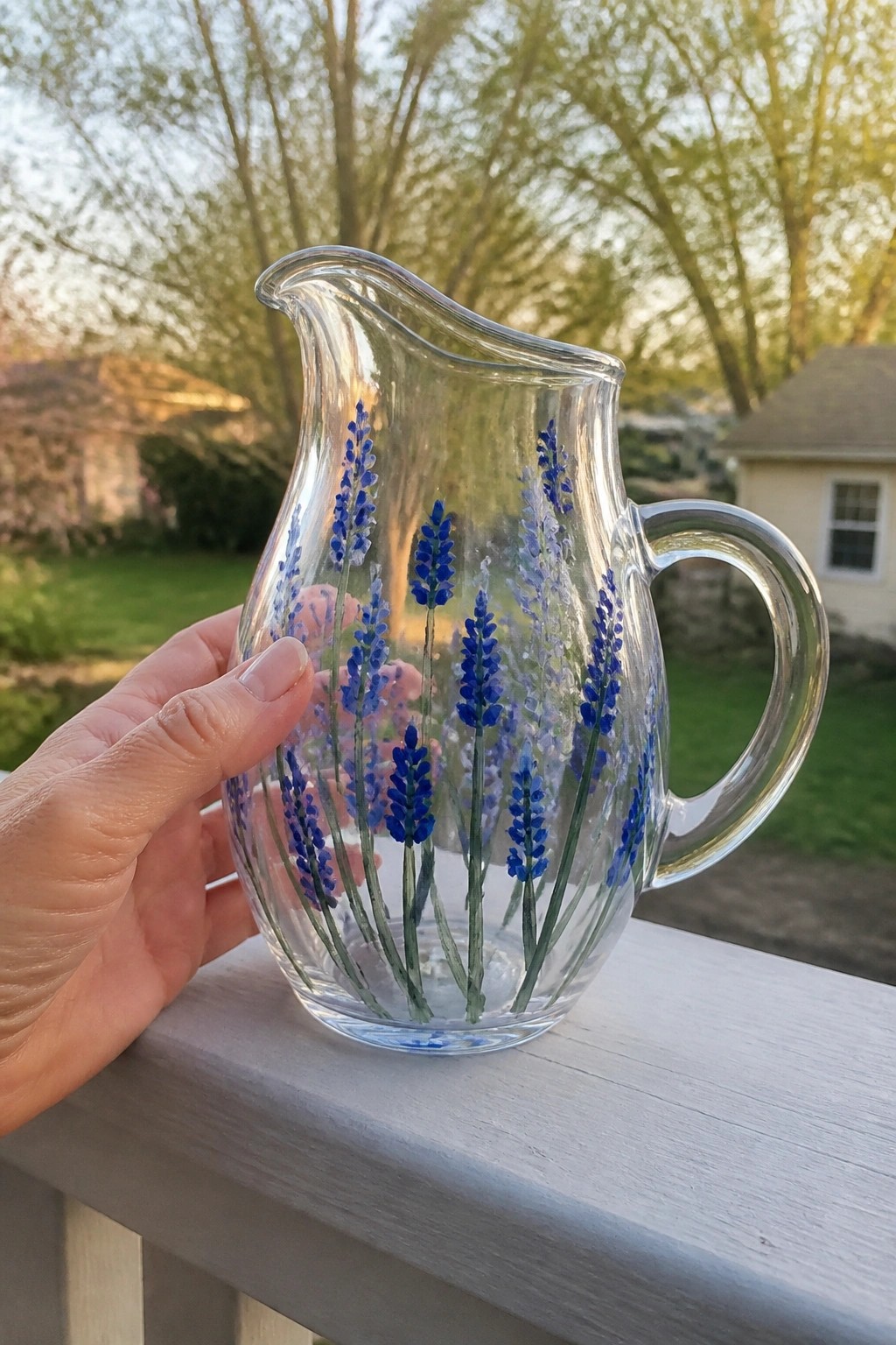 Hand holding glass pitcher painted with blue lavender flowers on white railing