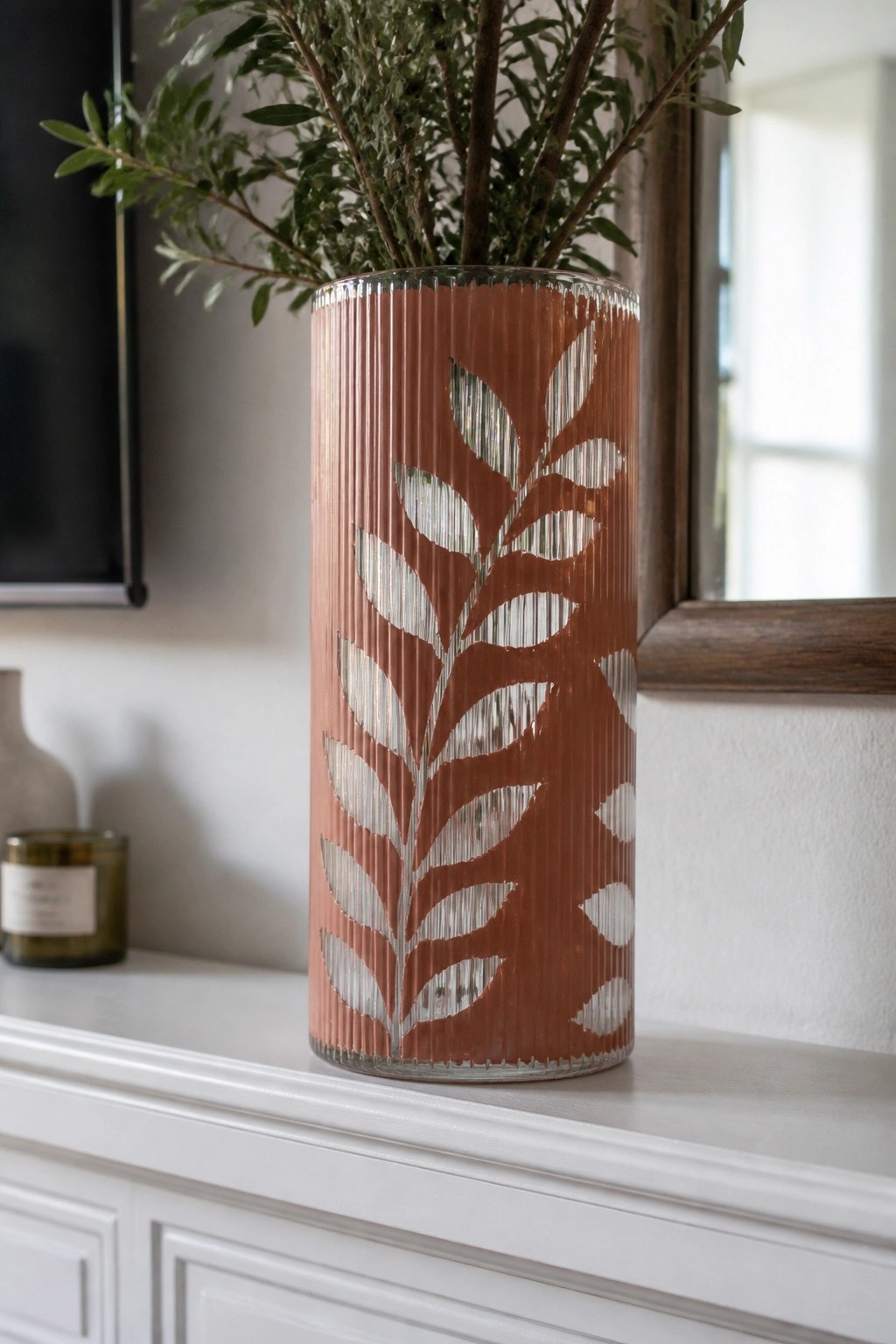 Terracotta ribbed vase with white leaf pattern holding green branches on mantel.