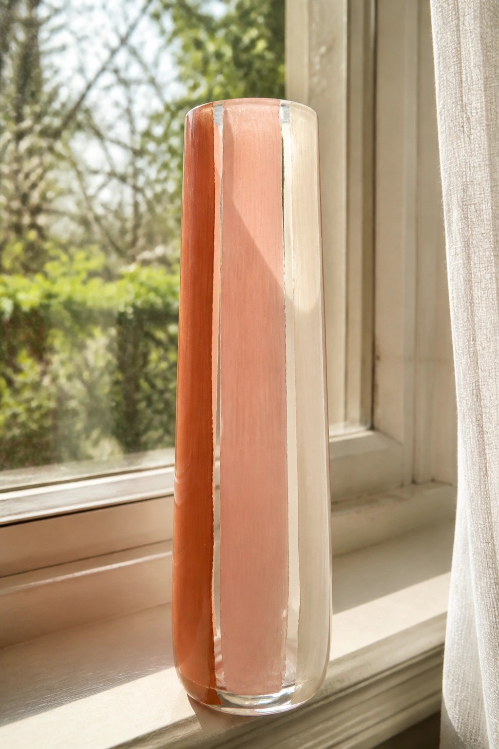 Tall striped glass vase in orange and pink on sunlit windowsill with trees outside