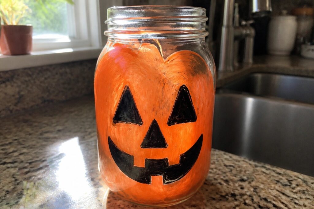 19 Easy Halloween Glass Painting Ideas Using Simple Silhouette Designs A clear glass mason jar painted orange with a black jack-o'-lantern face featuring triangular eyes and nose and a jagged mouth, sitting on a kitchen counter.