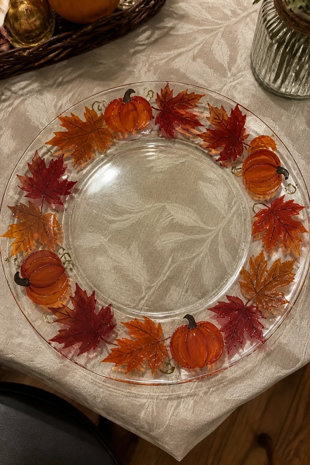 Clear glass plate rimmed with orange pumpkins and red maple leaves on white cloth