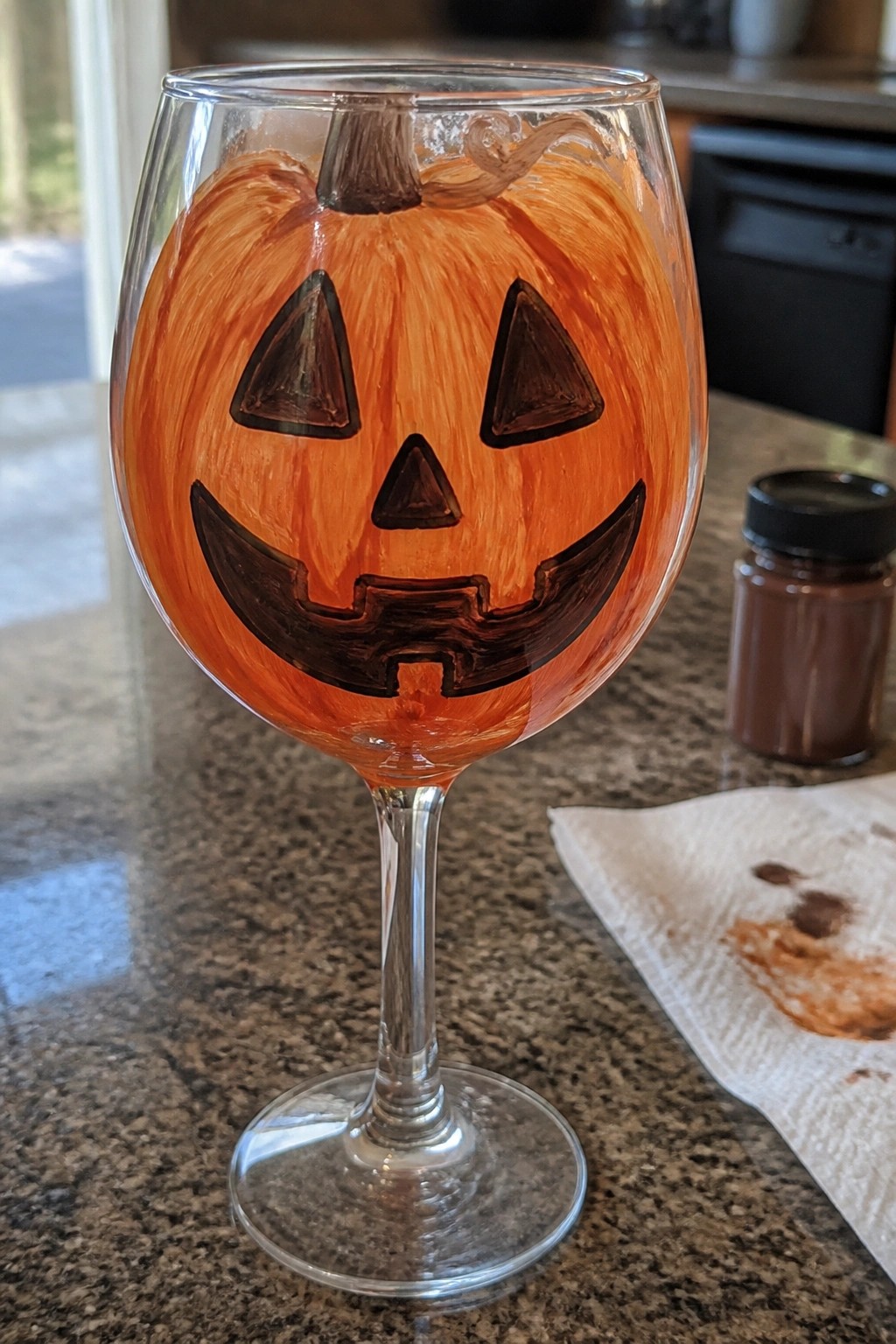 Wine glass hand-painted as orange jack-o'-lantern with black eyes, nose, and grin on counter