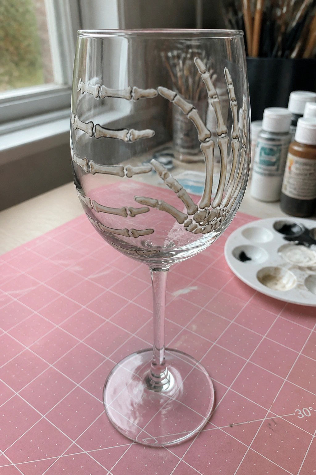 Skeleton hand bones painted on clear wine glass on pink gridded mat with supplies.
