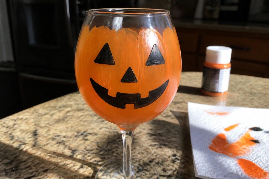 19 Easy Halloween Wine Glass Painting Projects for Beginners Wine glass painted orange as jack-o'-lantern with black face on kitchen counter