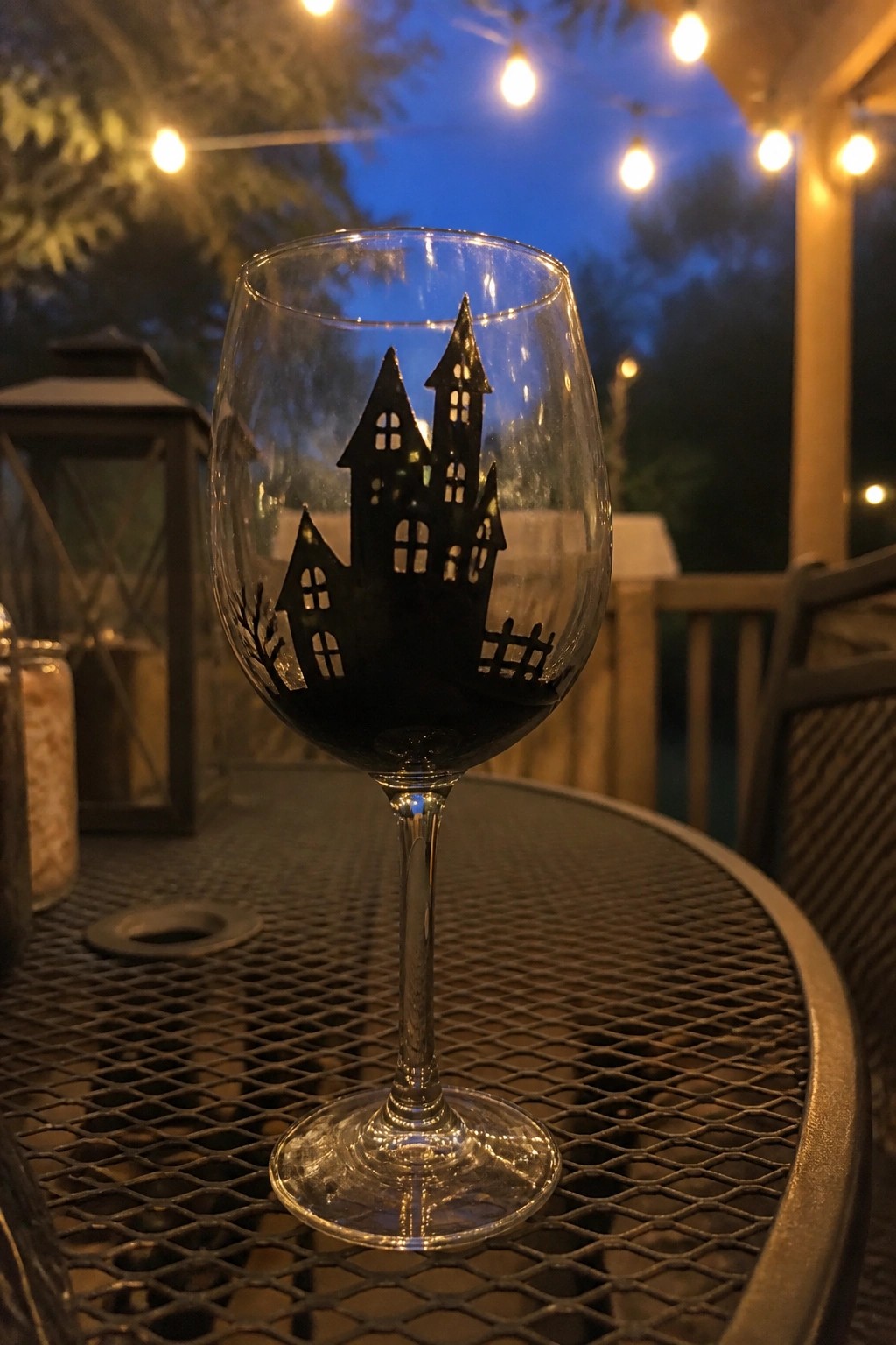 Wine glass with black haunted house silhouette filled with red wine on mesh patio table at night