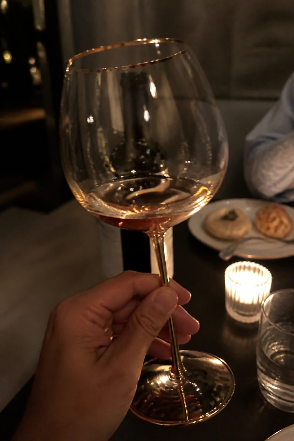 Hand holding gold-rimmed wine glass with rose wine above dimly lit table
