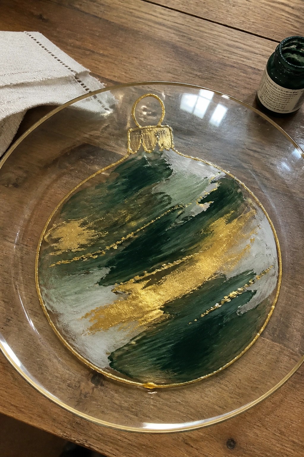 Gold-rimmed glass plate with abstract green, white, and gold Christmas ornament on wood