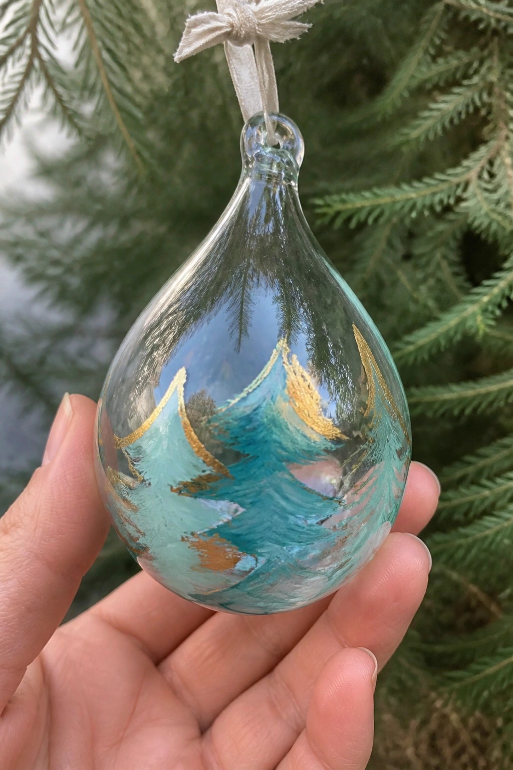 Hand holding teardrop glass ornament with teal waves and gold accents.