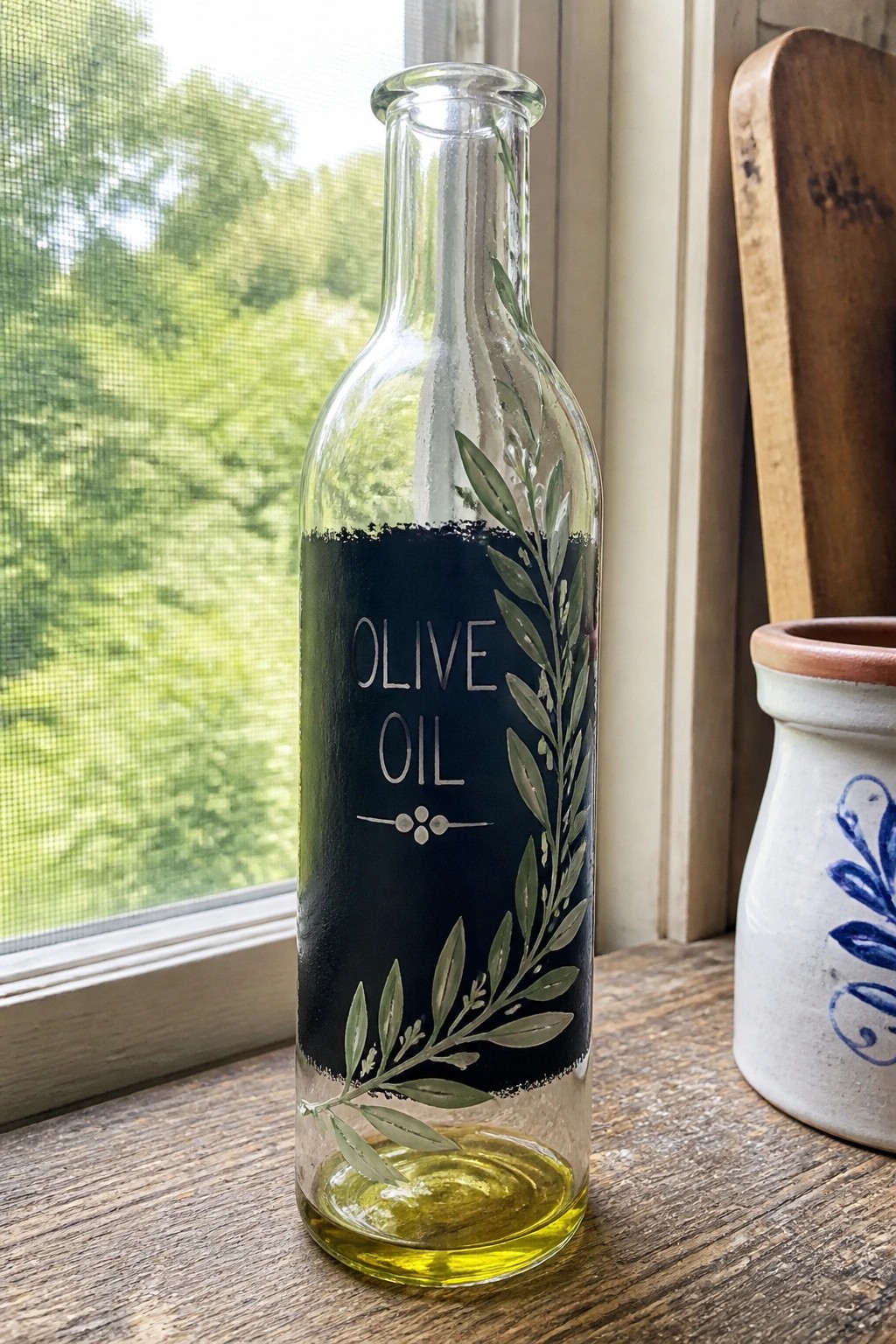 Glass olive oil bottle with black label and green branch designs on windowsill