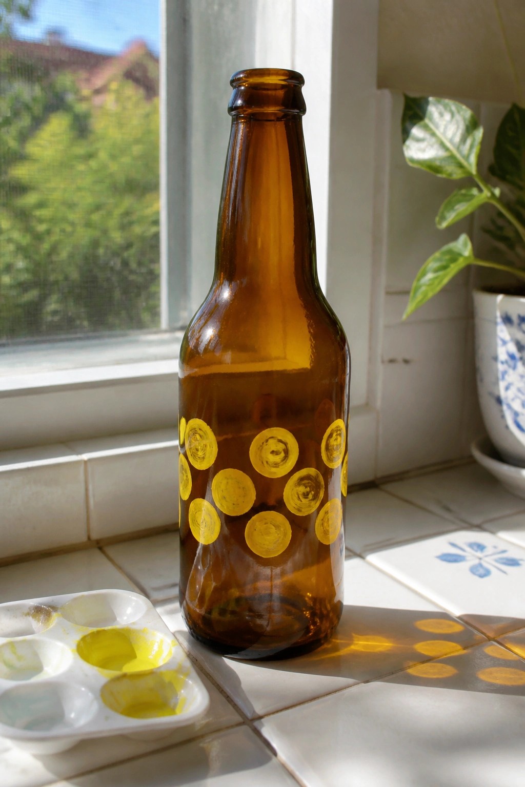 Brown bottle with yellow circles beside paint palette on sunny windowsill.