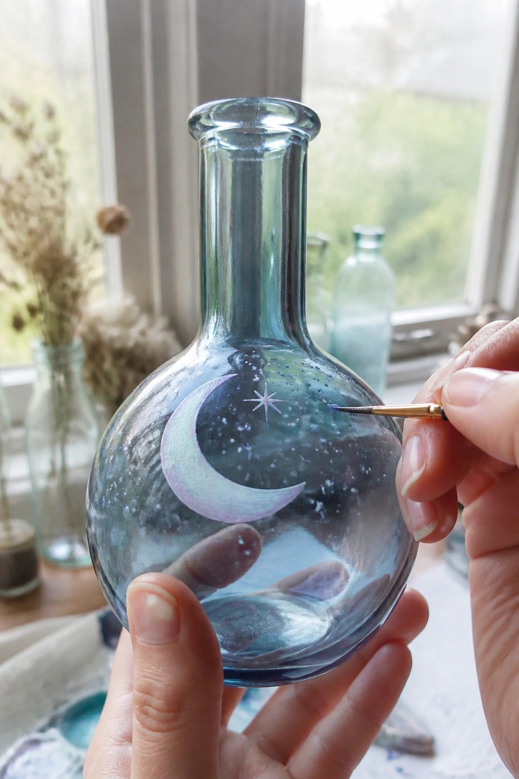 Hands painting a crescent moon and stars on a round glass bottle with a brush.