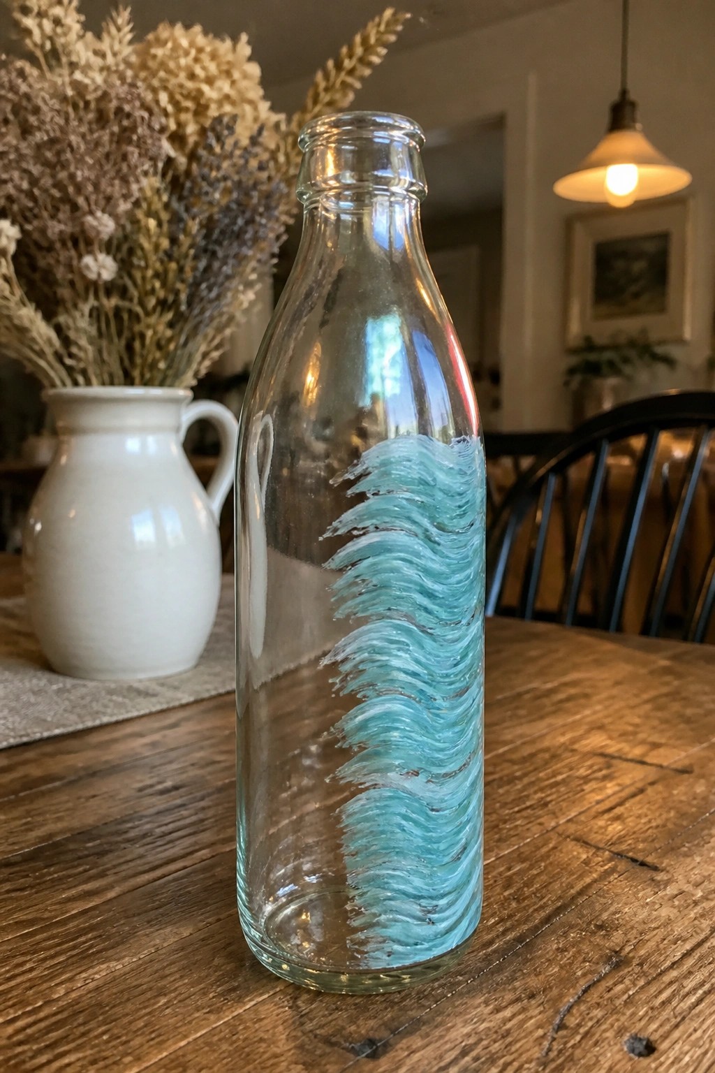 Clear glass bottle with hand-painted turquoise waves on wooden table beside white pitcher.