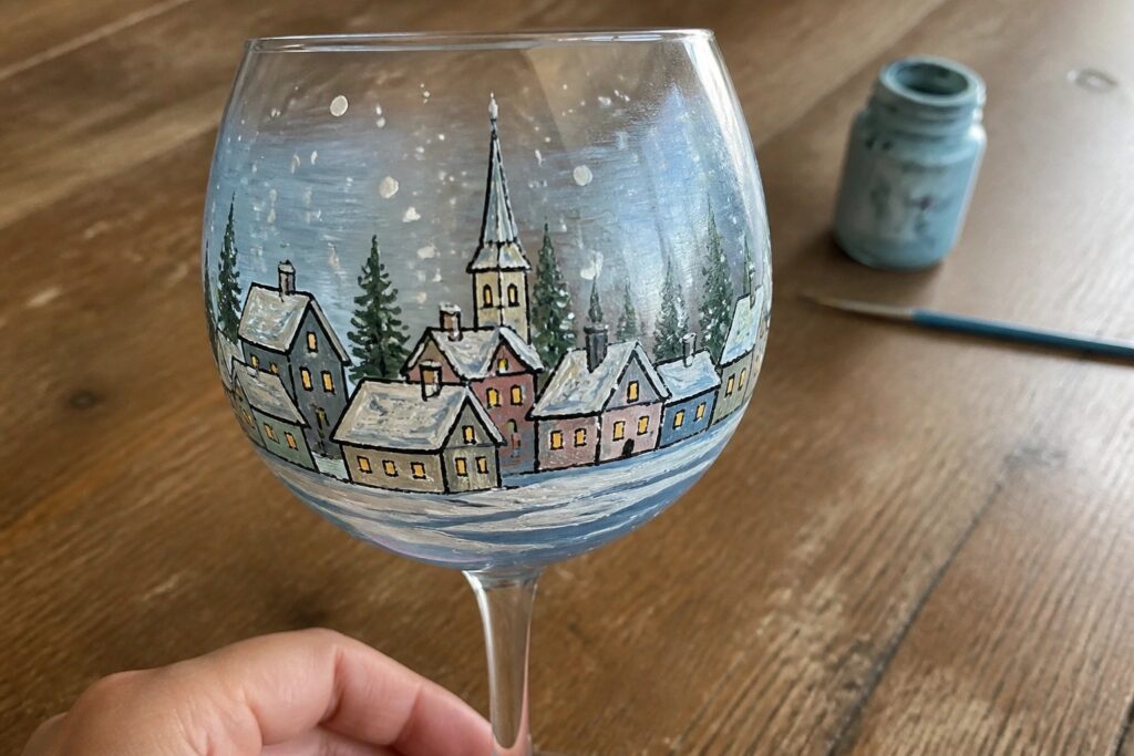 Hand-painted wine glass with snowy village scene of pastel houses and church on wooden table