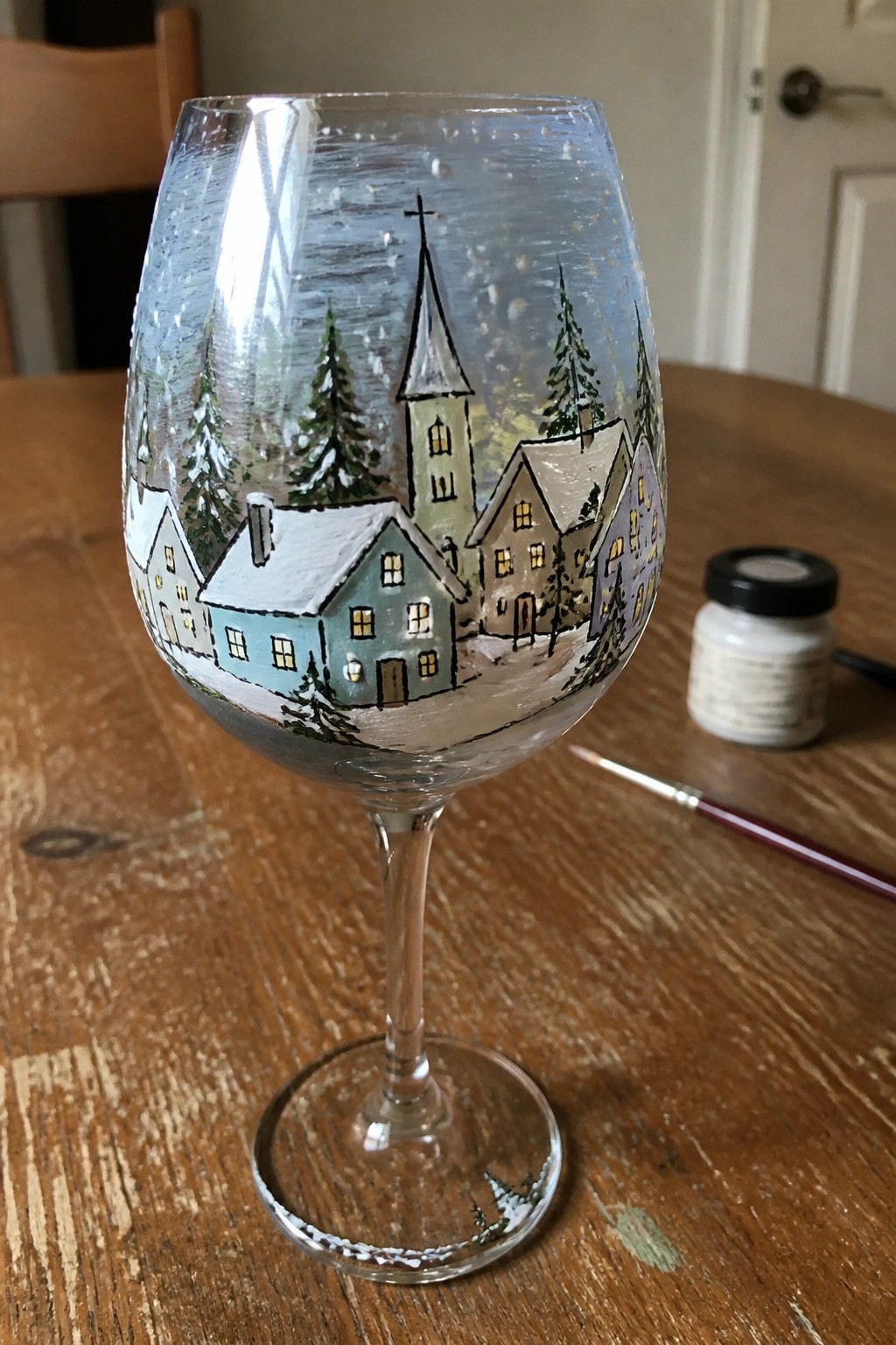 Hand-painted wine glass with snowy village scene of pastel houses and church on wooden table