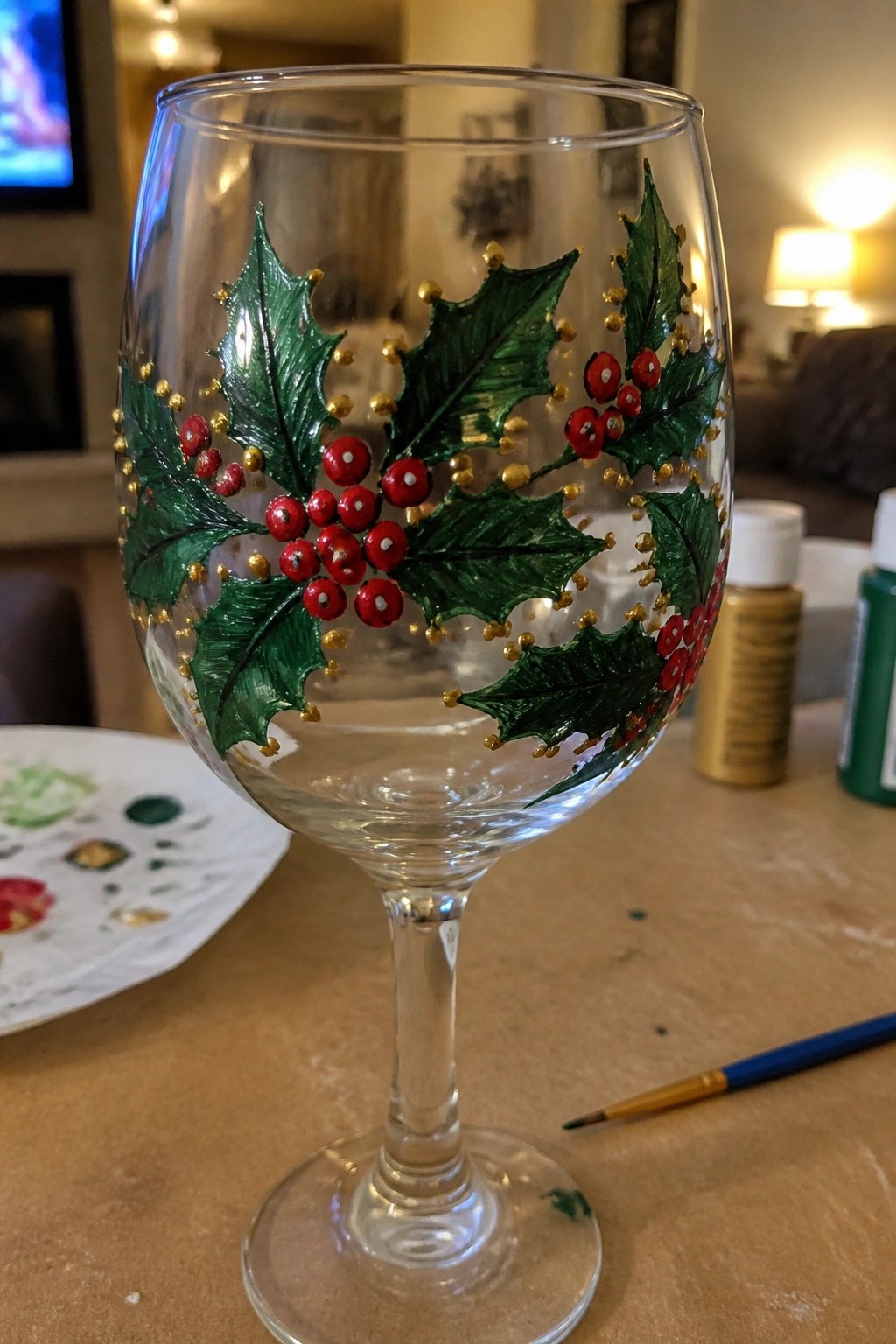Hand-painted wine glass with green holly leaves, red berries, and gold dots on table
