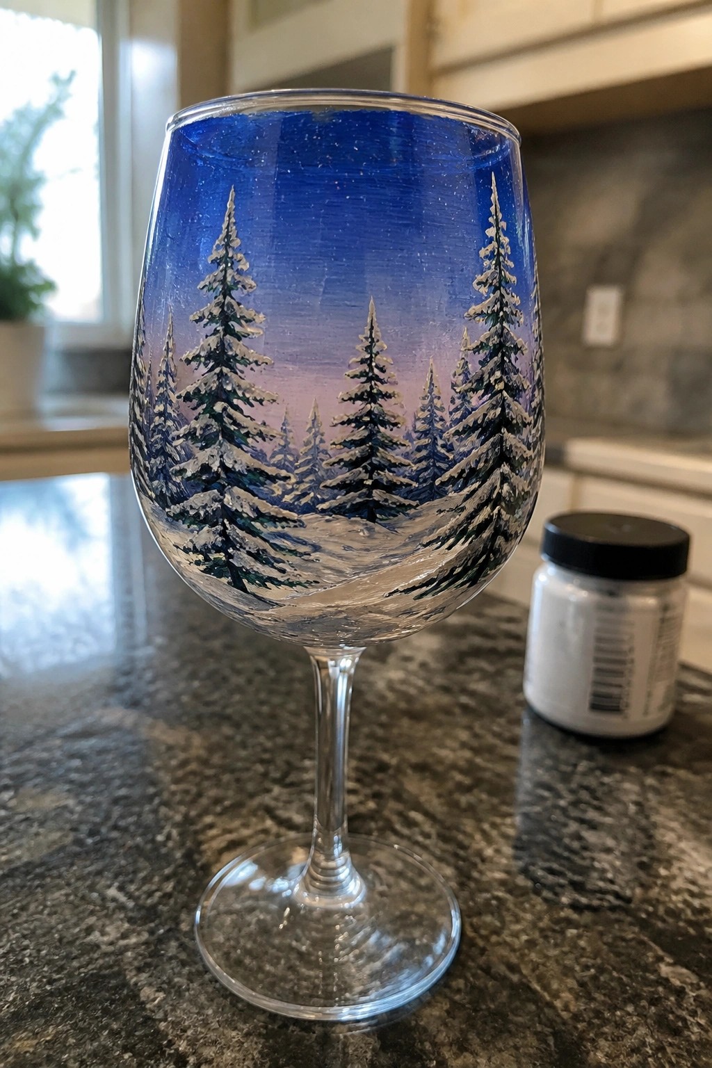 A clear wine glass painted with snow-covered pine trees against a blue-to-purple gradient sky rests on a dark countertop.