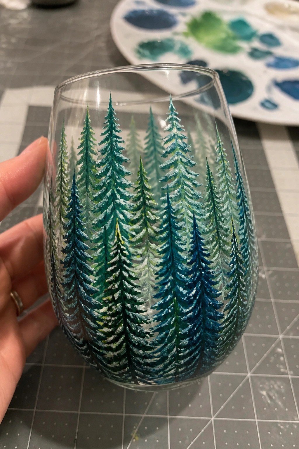 Hand holding stemless glass painted with blue-green pine trees, palette nearby.