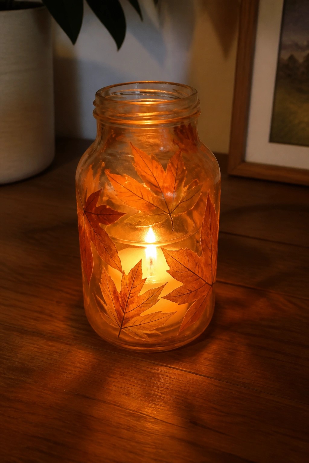 Glowing candle in glass jar decorated with pressed autumn maple leaves on wood.