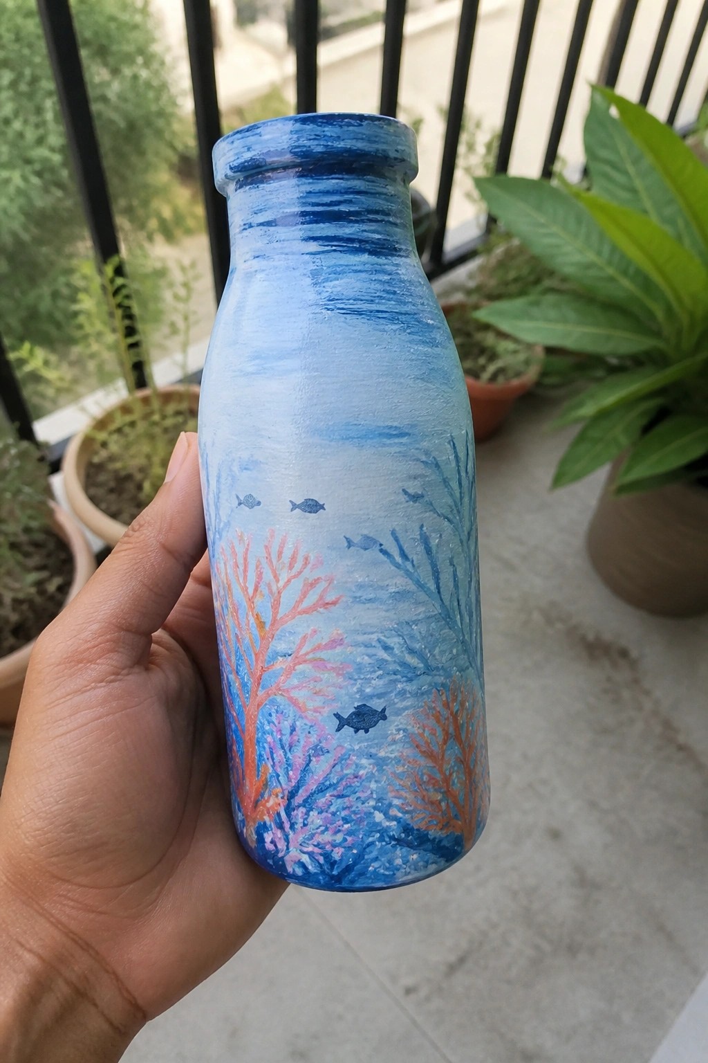 Hand holding blue painted bottle with colorful coral reefs and swimming fish.