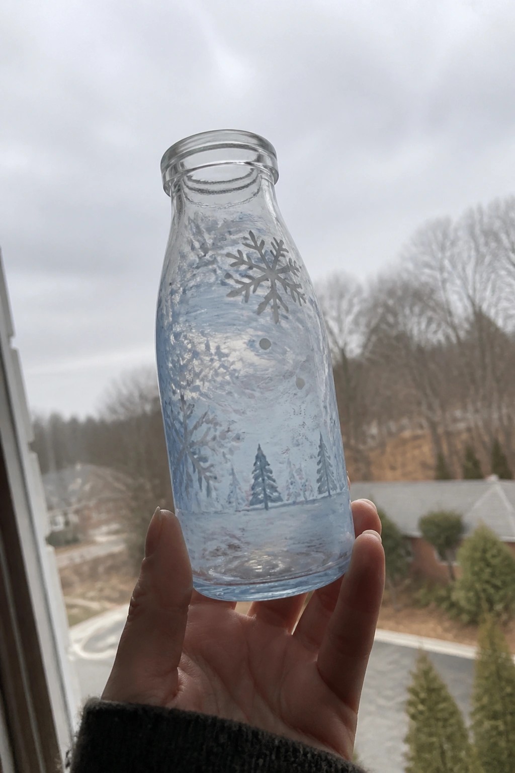 Hand holding painted glass bottle with snowflakes, trees, and snowy landscape.