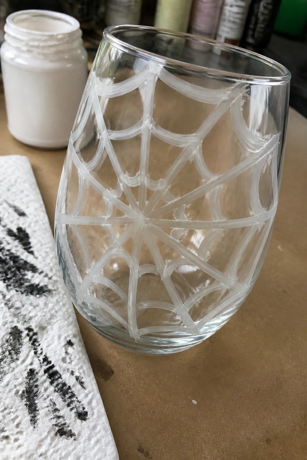 Stemless glass with white spiderweb paint design beside open jar and paper towel on wood.