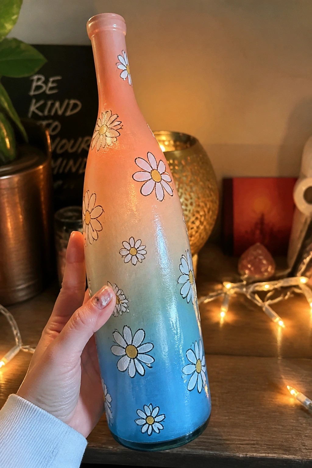 A hand holds a tall glass bottle painted with a pink to blue gradient and scattered white daisies with yellow centers.
