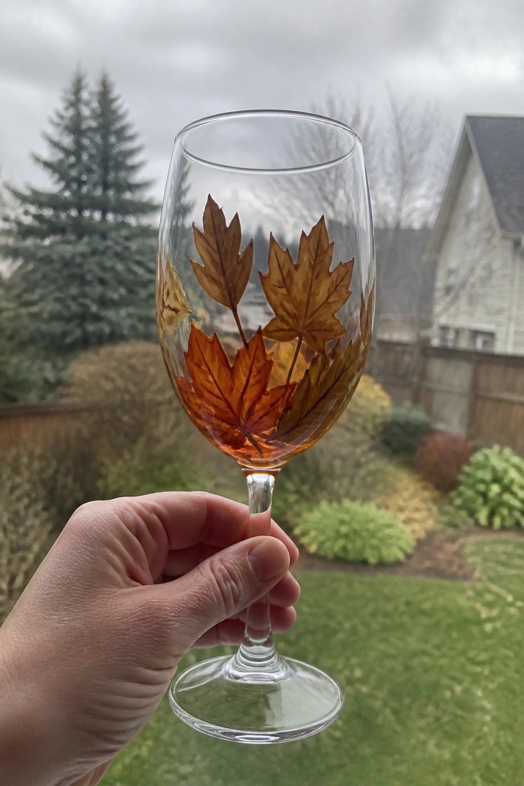 Hand holding wine glass painted with orange and brown autumn maple leaves outdoors.