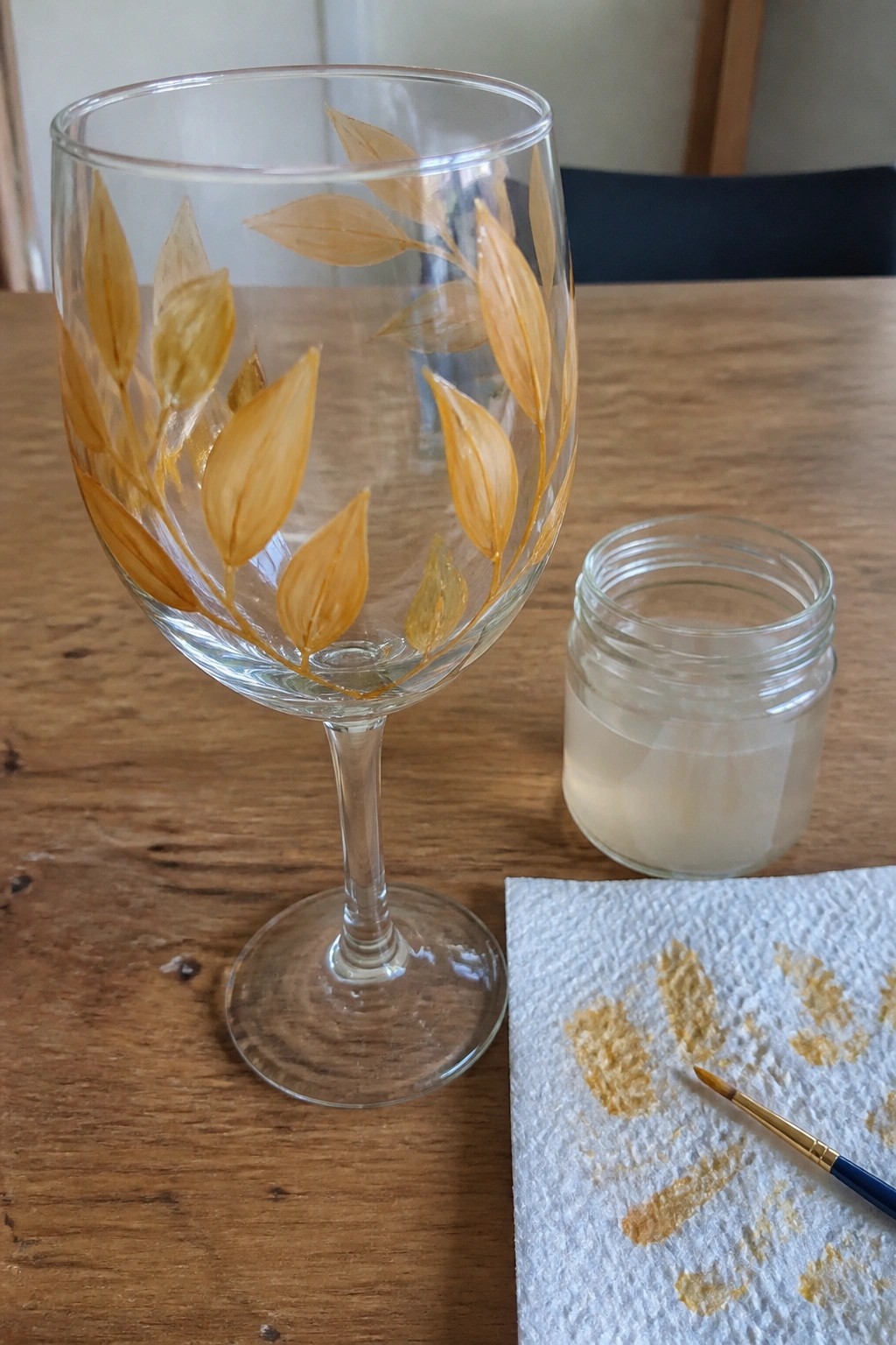 Wine glass with golden leaf designs beside paint jar, brush, and stained towel on table.