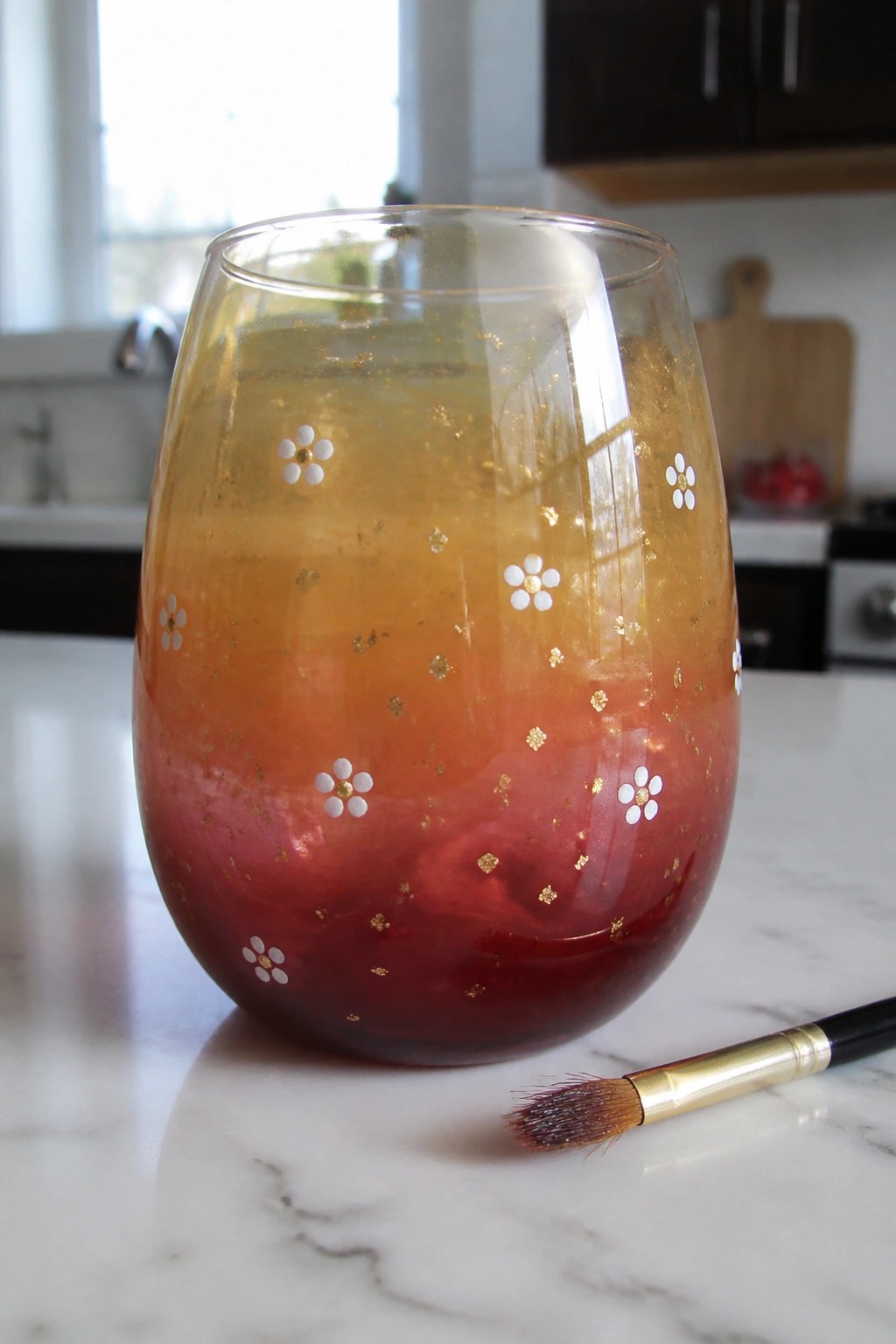 Hand-painted stemless glass with red-orange gradient, gold specks, white flowers beside brush