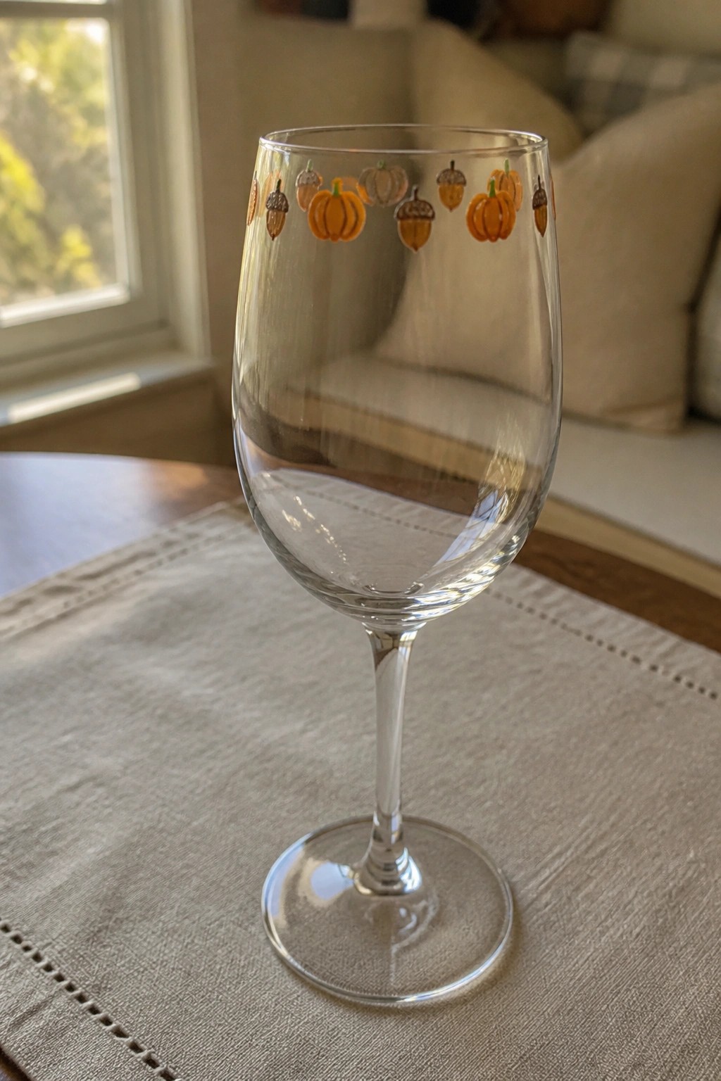 Clear stemmed wine glass with orange pumpkin and acorn designs on beige placemat.
