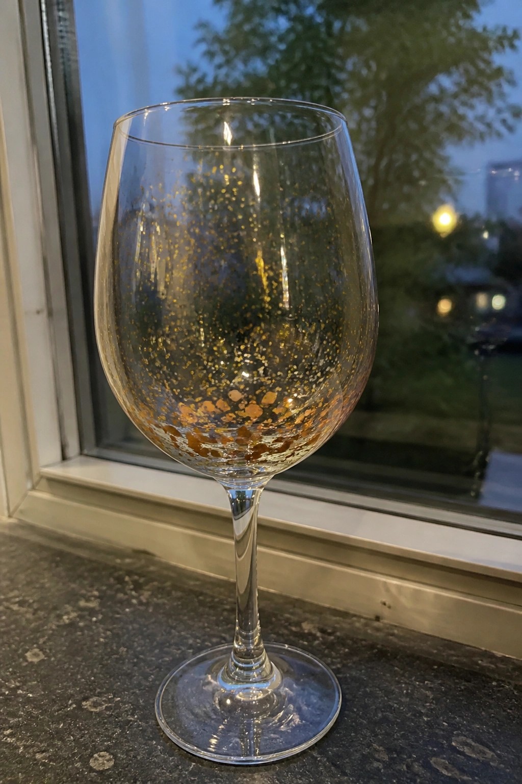 A clear stemmed wine glass with scattered gold and copper paint specks on the lower bowl rests on a dark surface in front of a window.