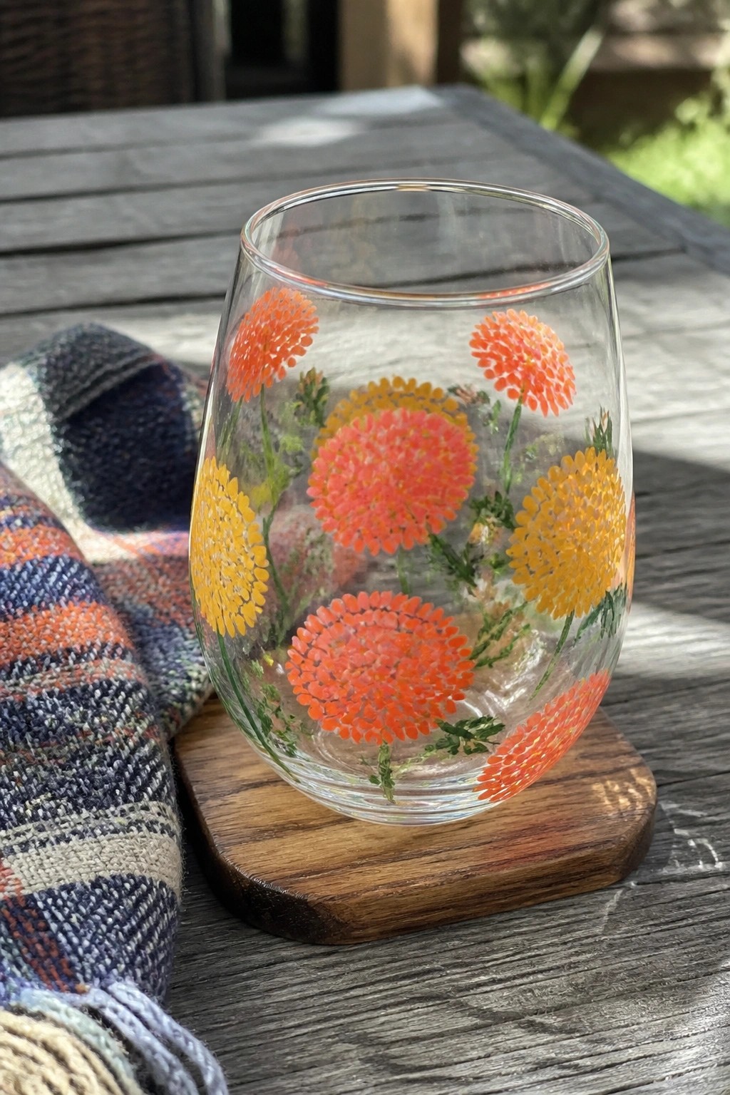 Stemless glass with orange and yellow flower designs on wooden table beside plaid cloth