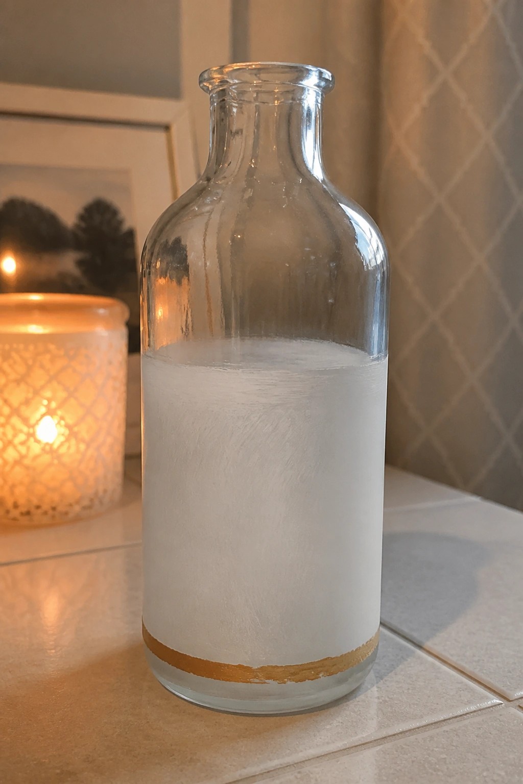 Glass bottle frosted white with gold band beside lit candle on tiles