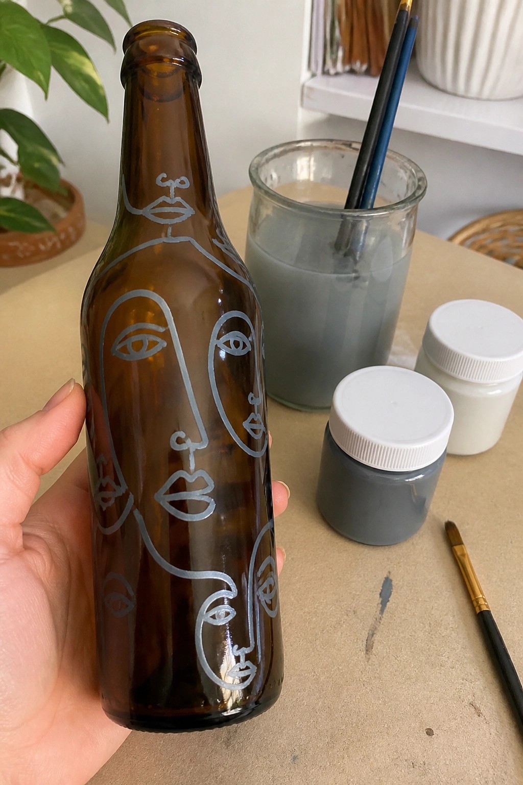 Hand holding brown bottle with white abstract face line art near paint jars and brushes