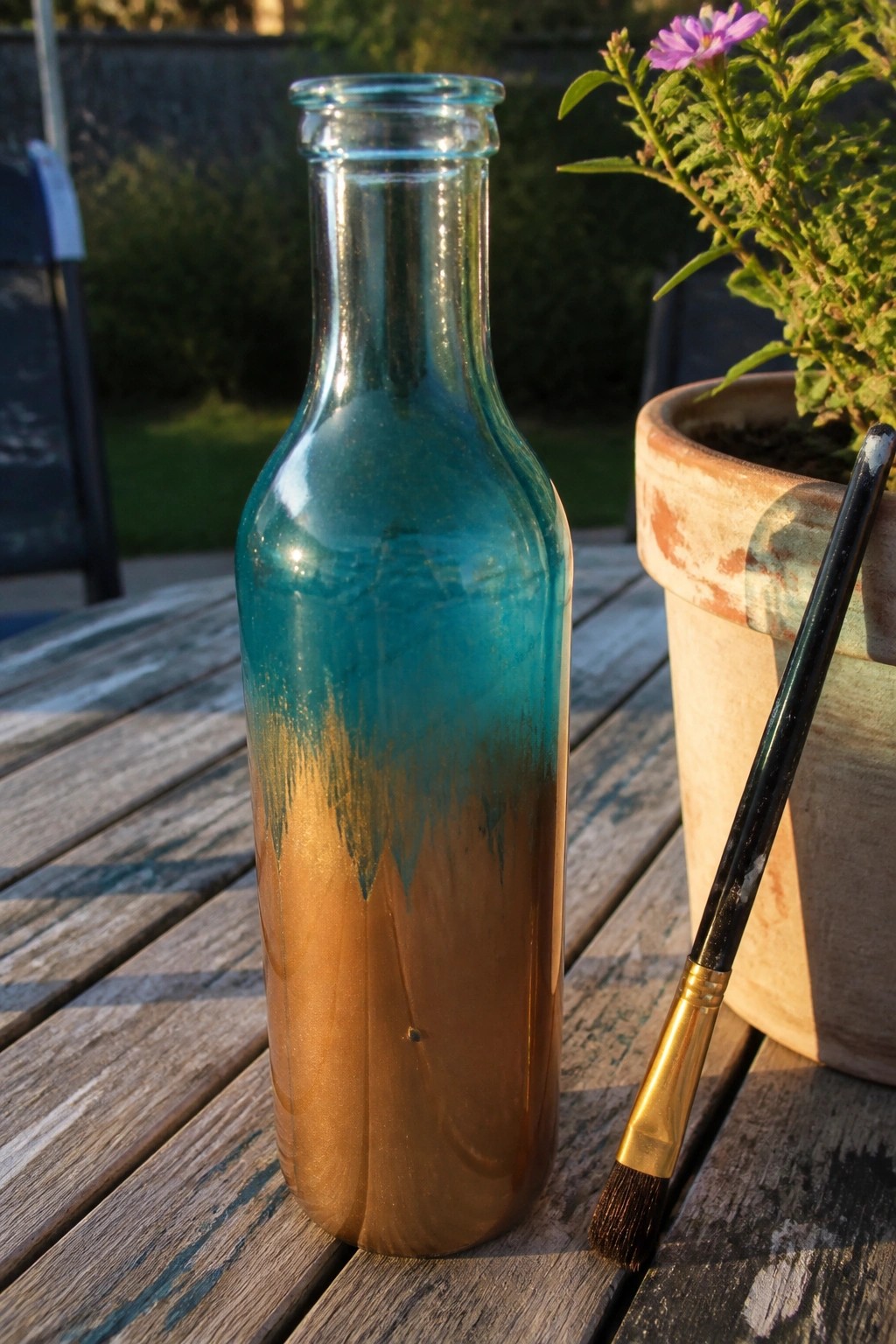 Turquoise and copper painted glass bottle with paintbrush near potted purple flower on wooden table