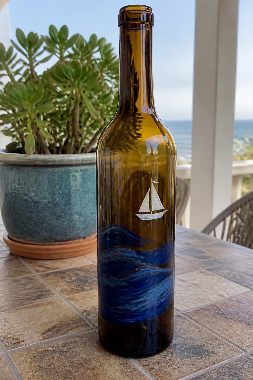 A brown glass bottle painted with blue waves and a white sailboat on a tiled table next to a potted plant.